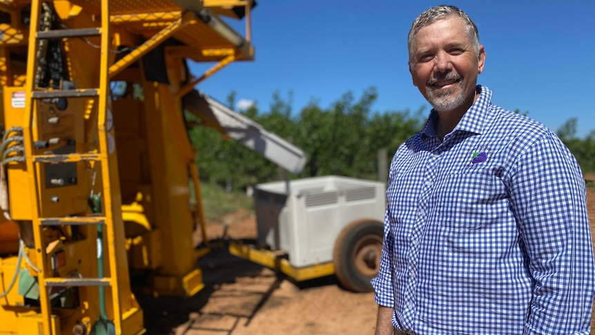 A man in a checked blue shirt standing in front of a fruit harvester.