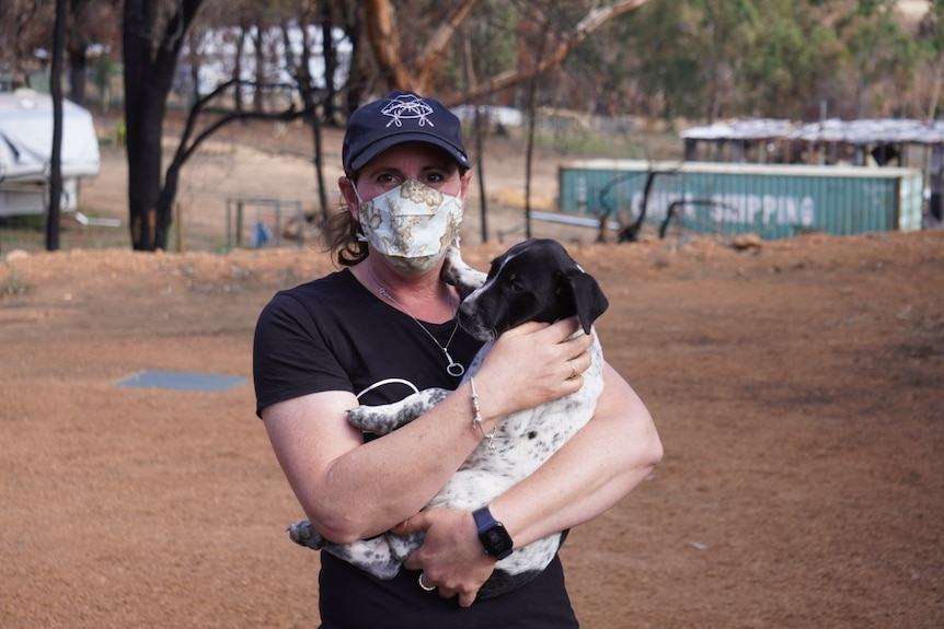Jessica wears a black shirt and white face mask, standing on a rural gravel driveway cradling a black and white puppy.
