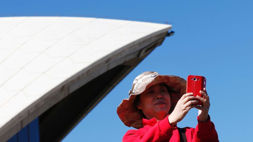 On a bright blue day, you see an Asian woman in bright pink take a selfie in front of one of the Sydney Opera House sails.