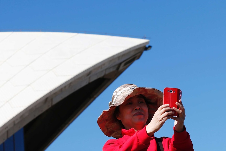 En un día azul brillante, ves a una mujer asiática vestida de rosa brillante tomando una selfie frente a una de las velas de la Ópera de Sydney.