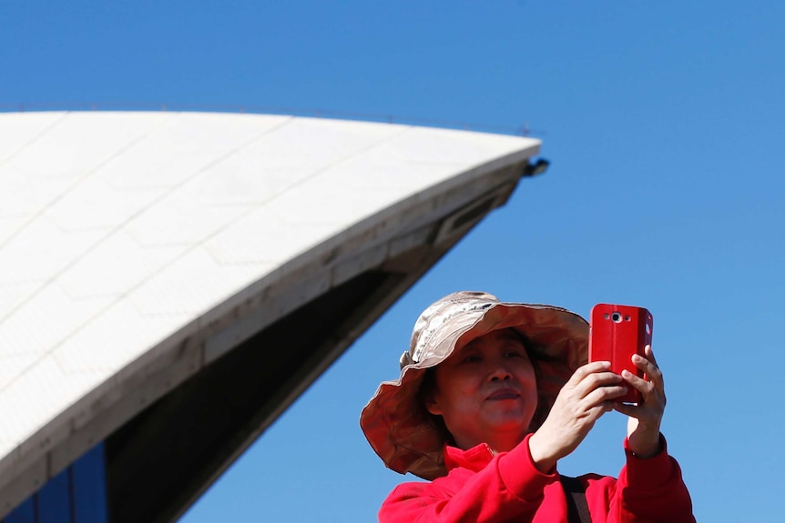 On a bright blue day, you see an Asian woman in bright pink take a selfie in front of one of the Sydney Opera House sails.