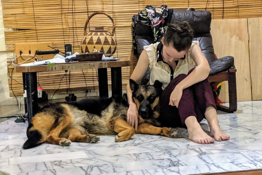 A young woman sits on the floor cuddling a lolling German Shepherd
