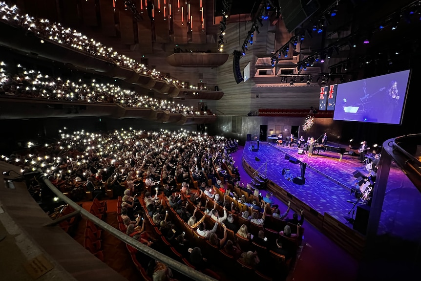 A crowd raise lit phones as Delta Goodrem performs on a stage bathed in purple light.