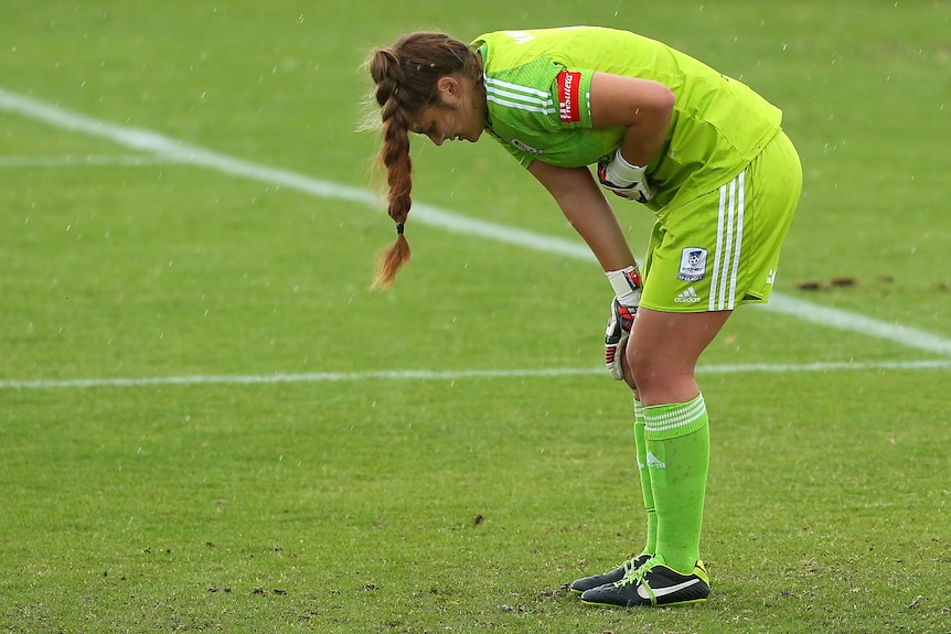 A soccer goalkeeper wearing light green leans over and puts her hand to her stomach