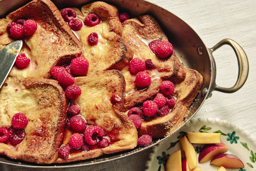 A baking tray filled with French toast, topped with maple syrup and raspberries. An easy brunch to prepare ahead of time.
