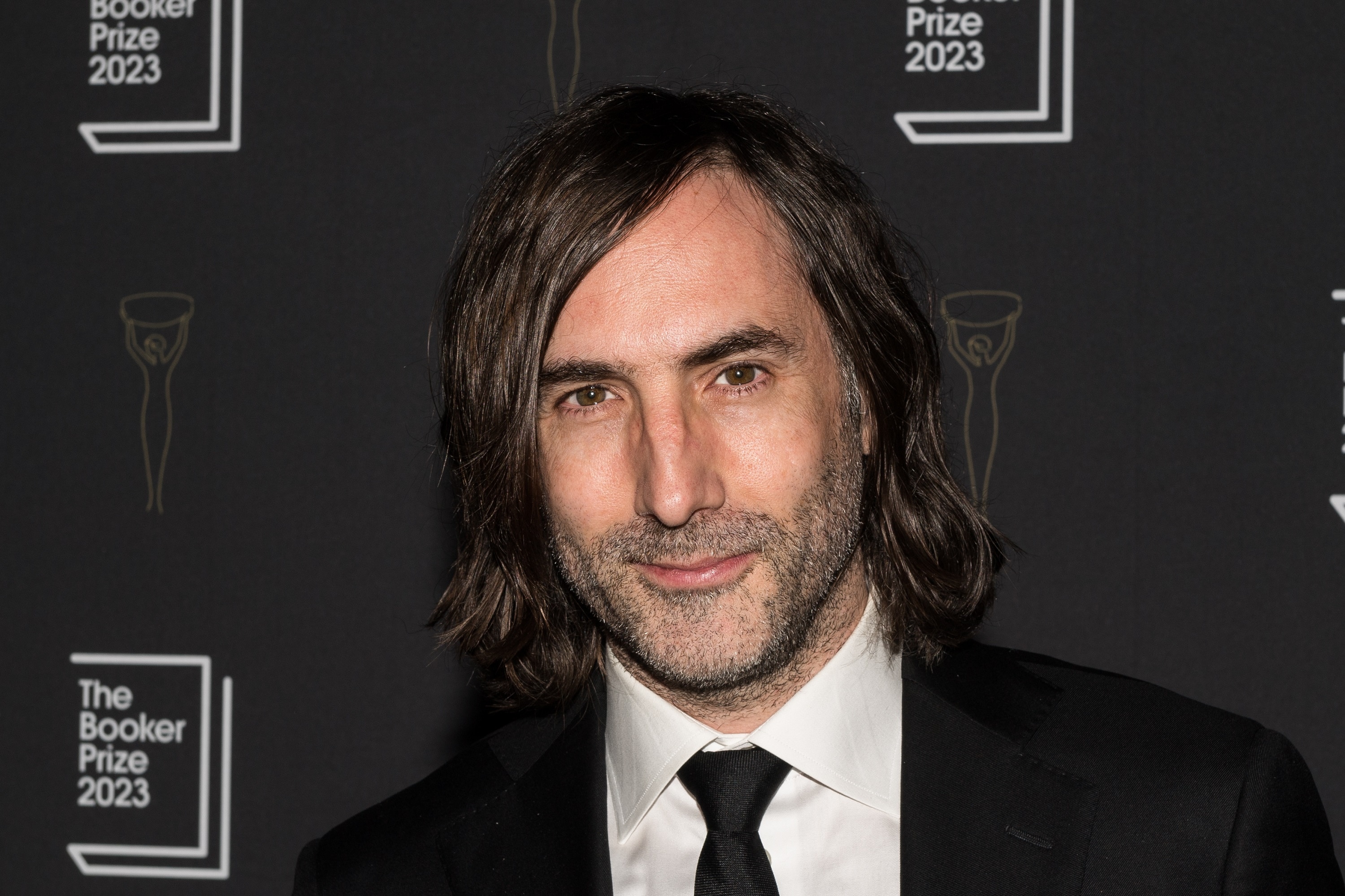 A middle-aged white man with chin-length dark hair, dark stubble and a smirk wear a suit and tie