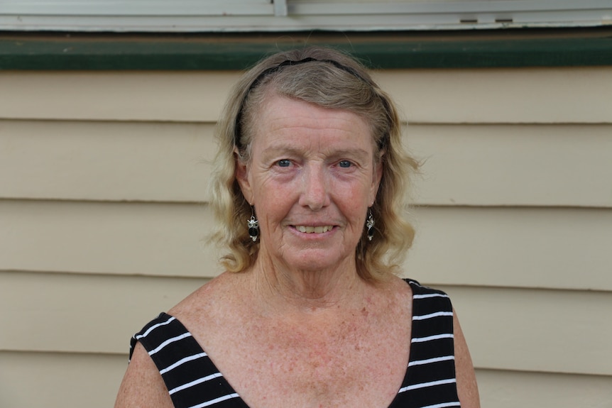 An elderly woman in a black and white striped dress stands in front of a cream wall of her house. 