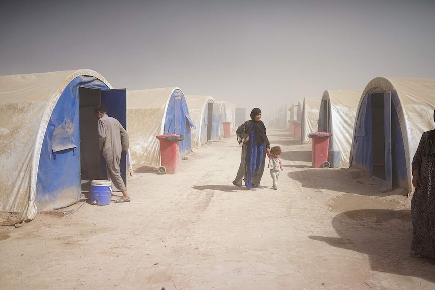 A mother and child wander between a row of tents in the windswept desert.