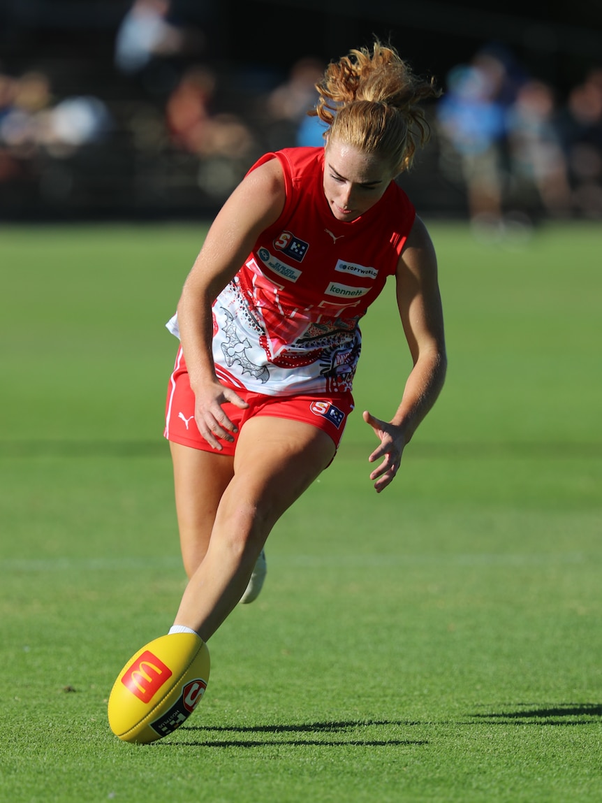 A woman playing Aussie rules in a red guernsey leans over to get a football.