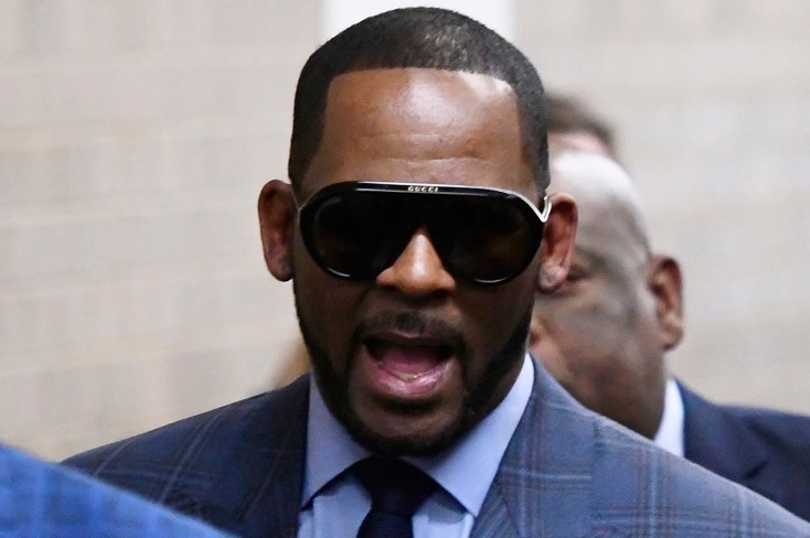 A man in a suit wearing sunglasses looks at the camera with his mouth open as he walks with a group