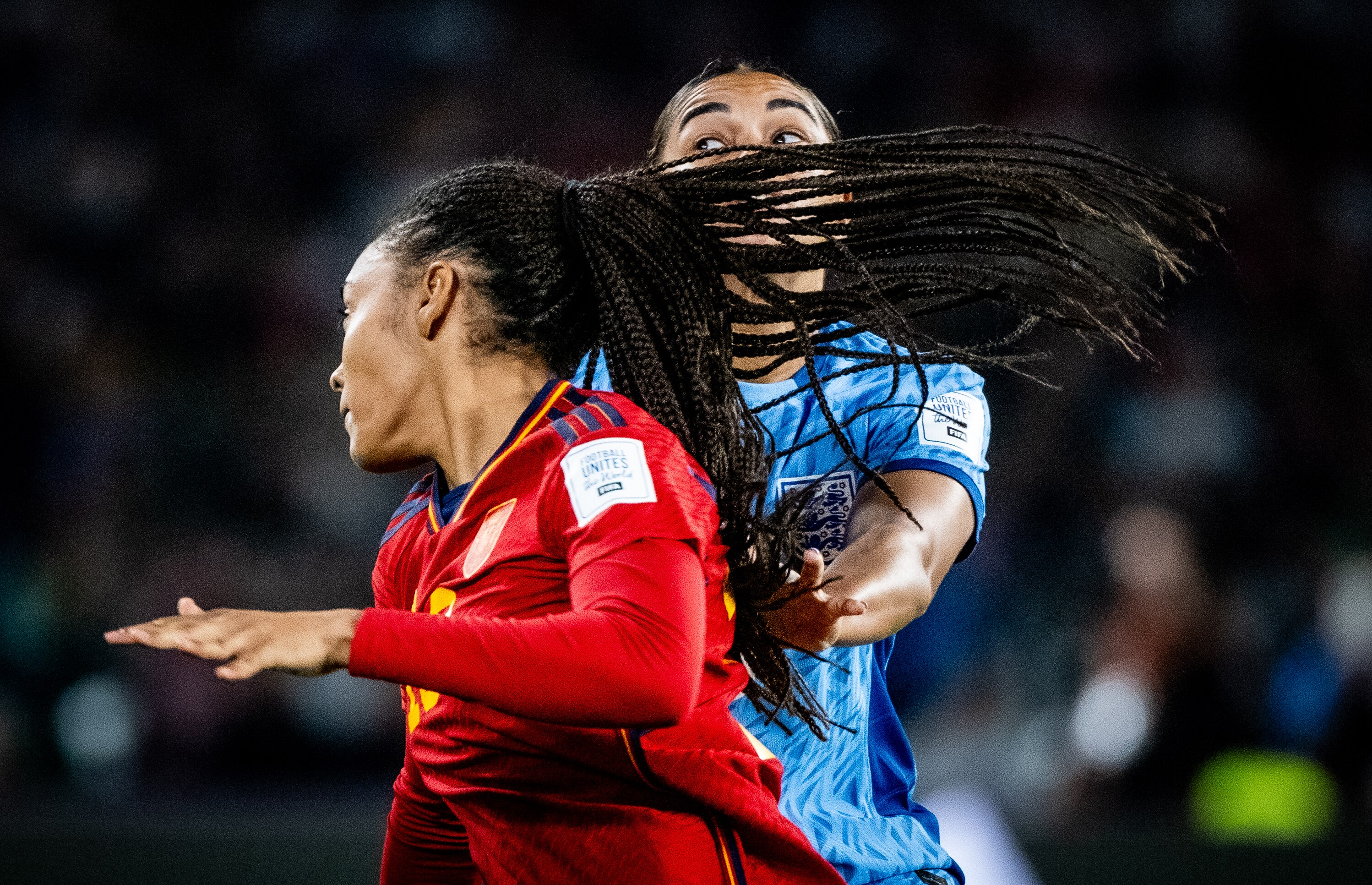 A female soccer players long hair blows across another woman's face and only her wide open eyes can be seen behind the hair.