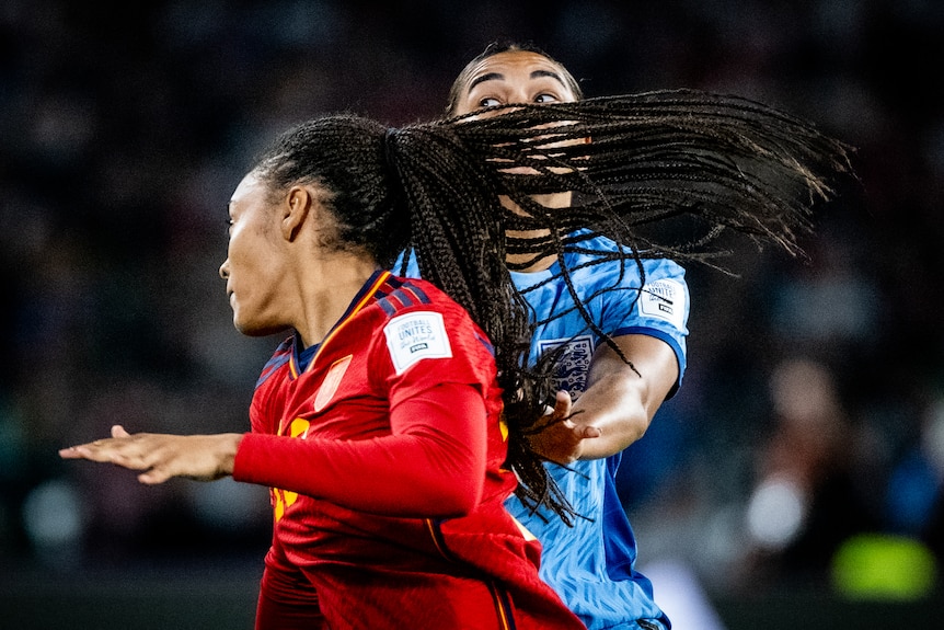A female soccer players long hair blows across another woman's face and only her wide open eyes can be seen behind the hair.