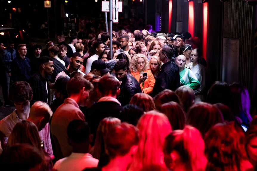 Tłum zbiera się przed nocnym klubem, z blondynką w pomarańczowym stroju w centrum uwagi.