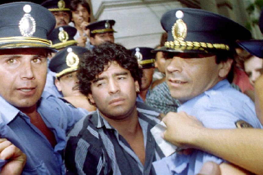 Diego Maradona looks dazed amid a throng of police officers.