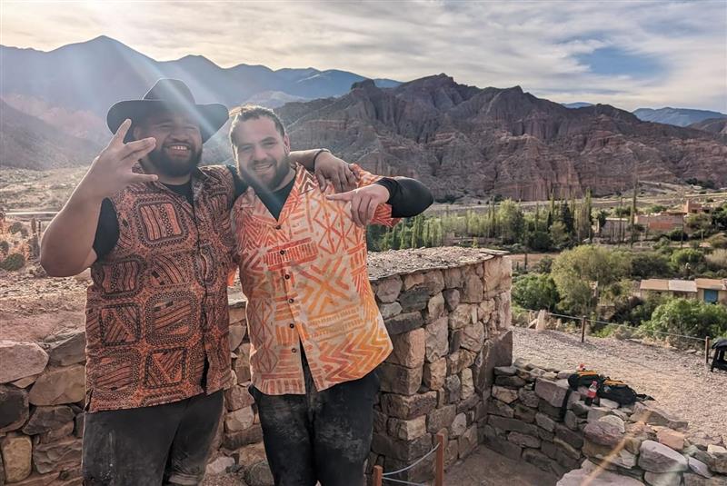 Two men stand outside wearing printed button up shirts in what looks like an desert type area. One of the men wears a cowboy hat