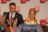 A man and woman stand smiling in front of a Labor banner. In the foreground are hands holding up phones taking photos 