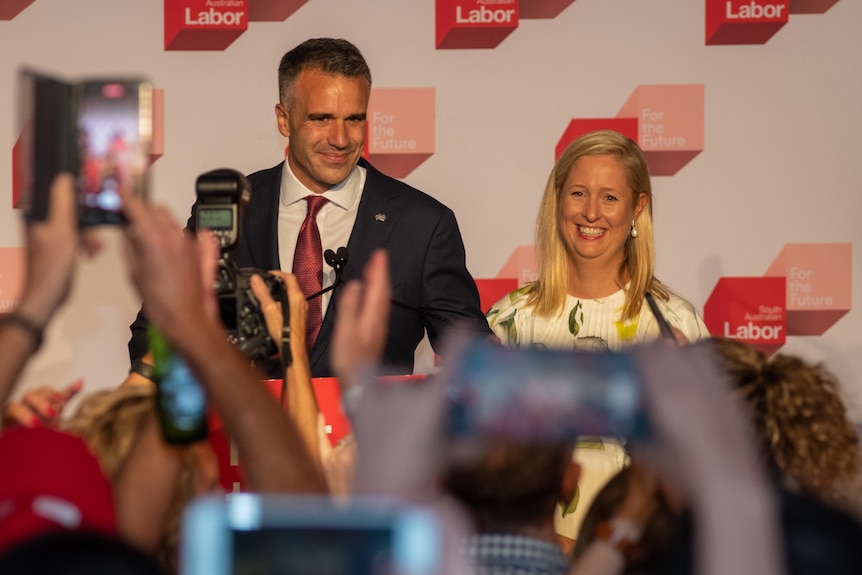 A man and woman stand smiling in front of a Labor banner. In the foreground are hands holding up phones taking photos 