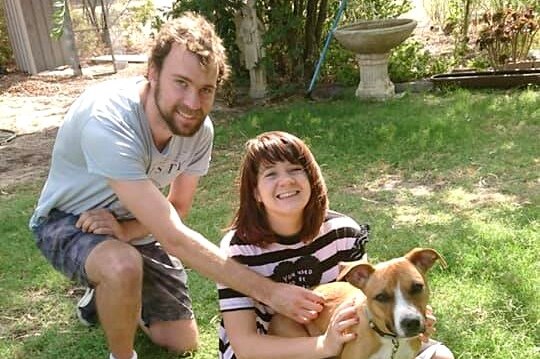 A , smiling young man kneels down next to a woman sitting on the grass holding a small brown dog.