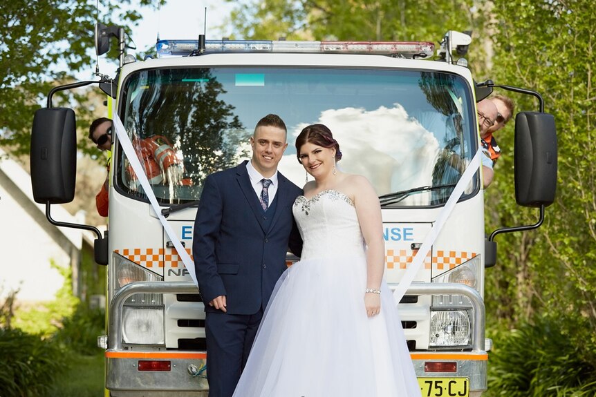 A bride and groom stand next to each other in front of a SES truck, with SES volunteers putting their heads out the windows.