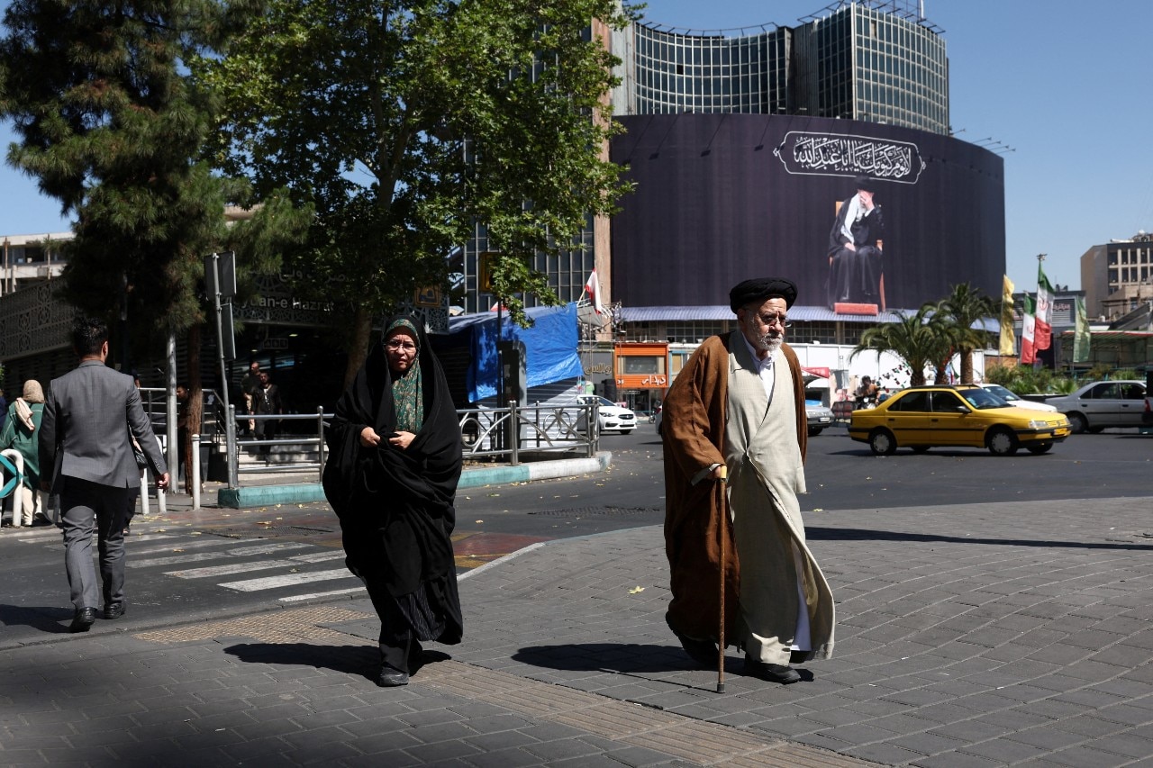 People walking near a busy street, looking on. Behind them a large billboard showing an elderly man sitting down can be seen.