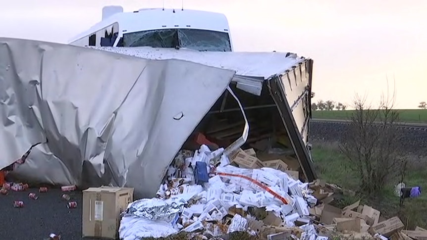 Cardboard boxes, cans and white papers spills out of the end of a busted trailer lying across the Western Highway.