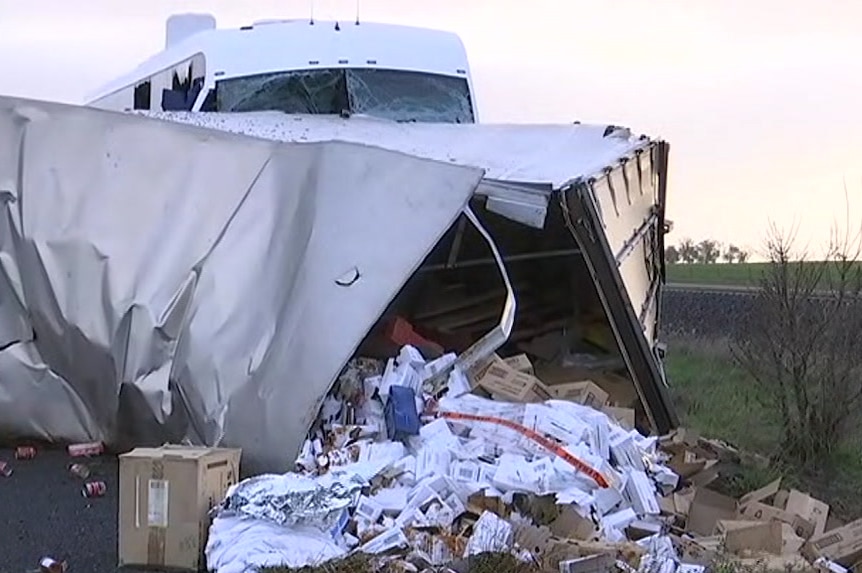 Cardboard boxes, cans and white papers spills out of the end of a busted trailer lying across the Western Highway.