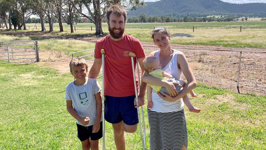 A man with one leg, on crutches, stands in front a paddock with a boy and woman holding a baby