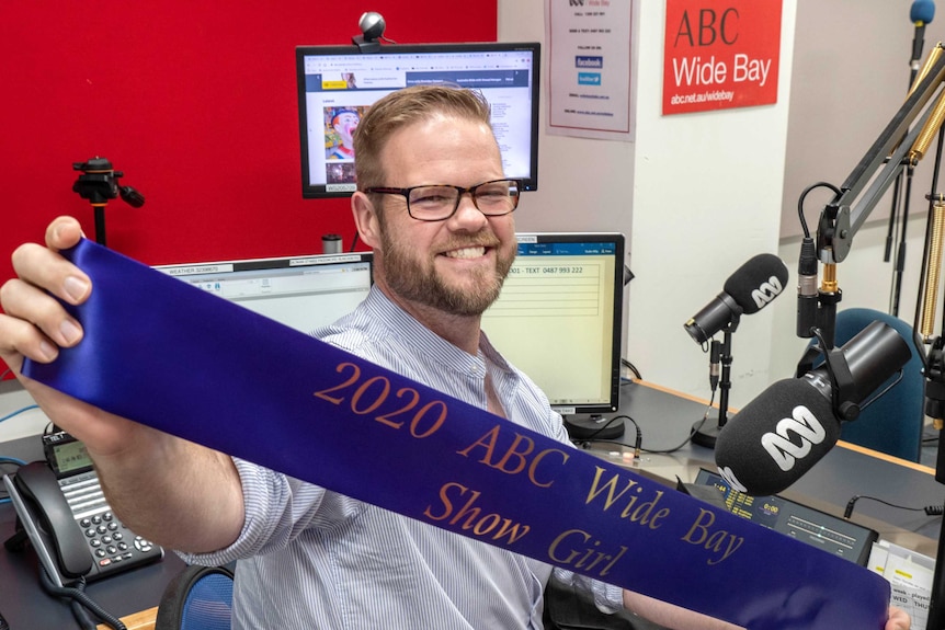 mid shot of a radio presenter sitting in the studio holding a purple sash that reads 2020 ABC Wide Bay showgirl