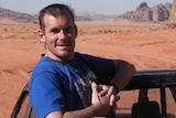 A man stands on the back of a car in a desert setting