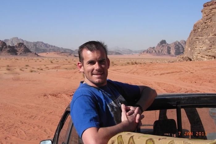 A man stands on the back of a car in a desert setting