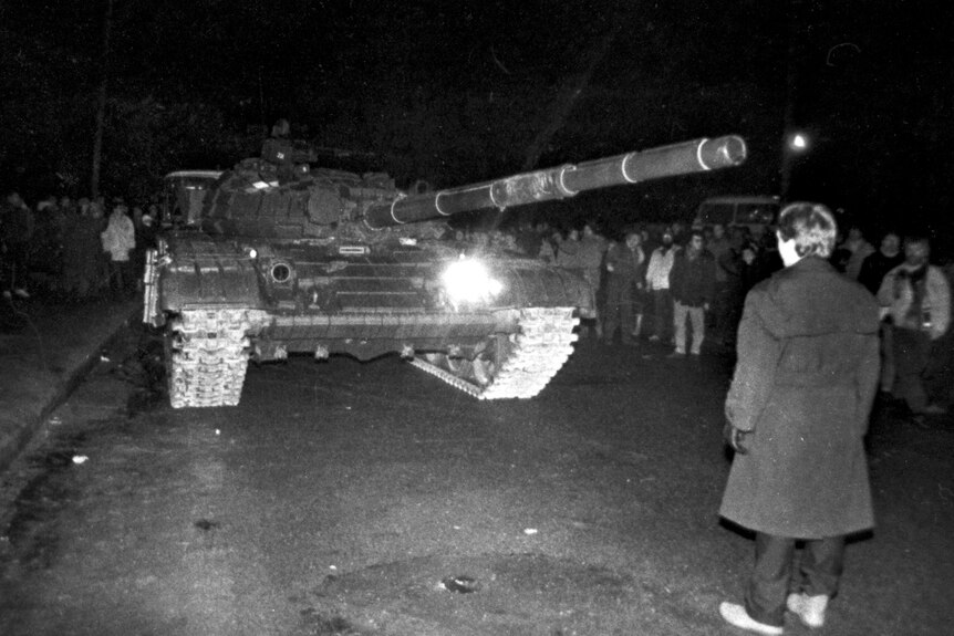 A black and white image. A man in a long coat stands in the middle of the road before a tank pointed at him