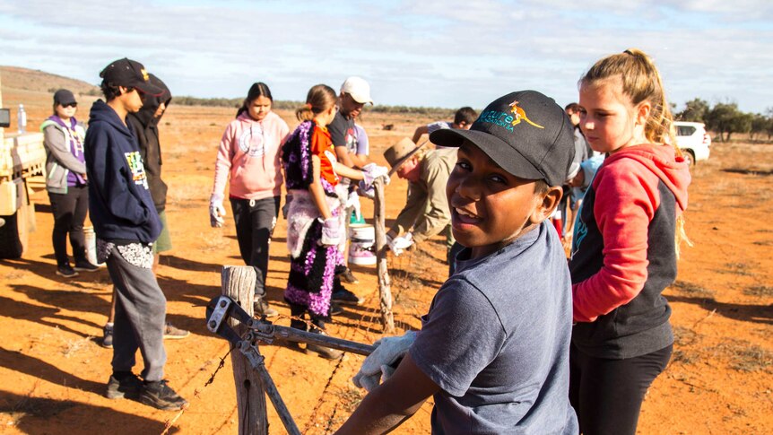 A young boy smiles at the camera in the foreground while using bolt cutters to sever barbed wire fencing. Children in background