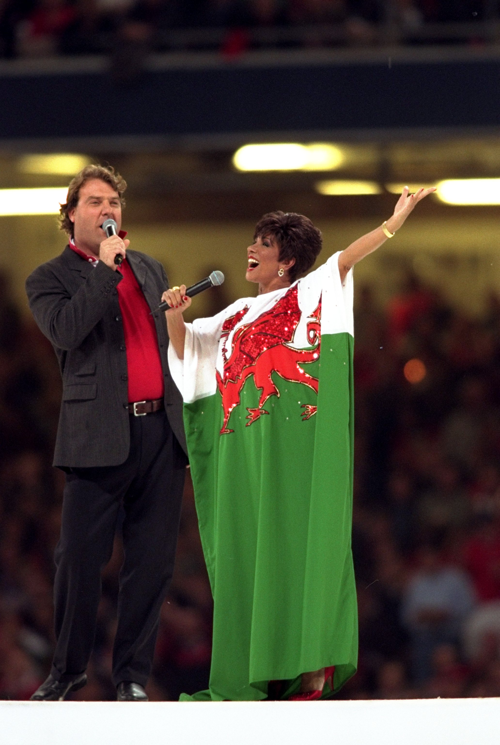 Bryn Terfel and Shirley Bassey perform on stage, Bassey in a kaftan dress of the Welsh flag, before a stadium audience.