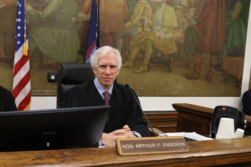 The judge sits at the bench. A large mural is behind him. His clerk is to his right. Both are looking at the camera.