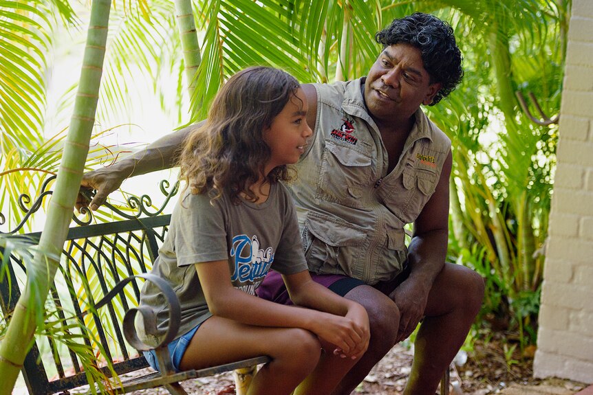 A young girl smiles as she talks to her dad. They are sitting on a bench stool in a tropical garden.