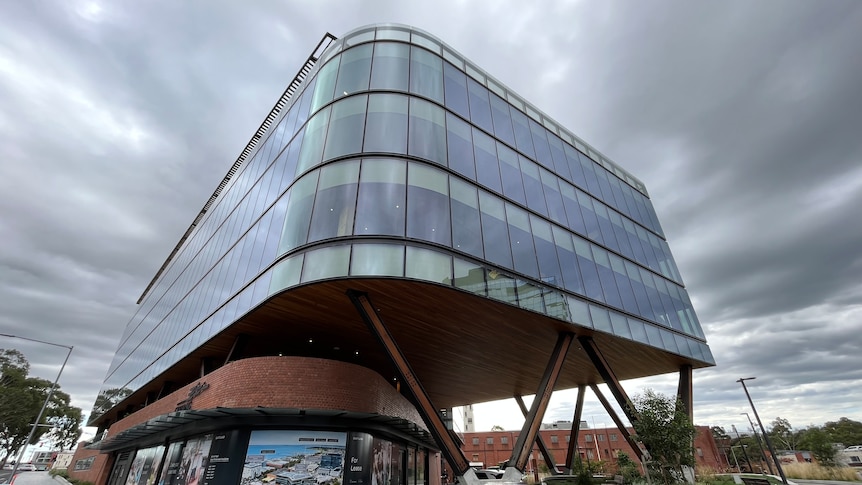 A four-storey building with large glass windows wrapping around it and brick for the first floor sits under a grey and dark sky.