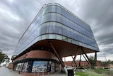 A four-storey building with large glass windows wrapping around it and brick for the first floor sits under a grey and dark sky.