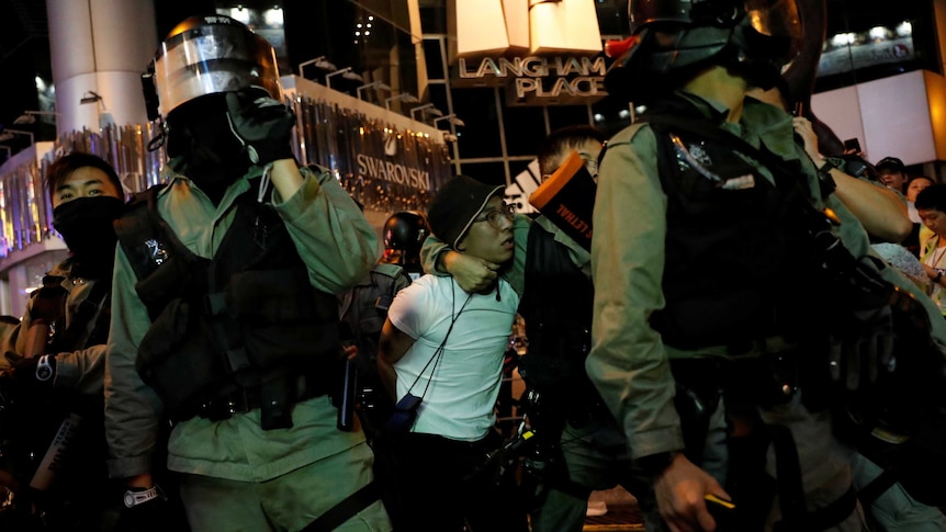 A protester in a white shirt and glasses is dragged away by a group of police in riot gear