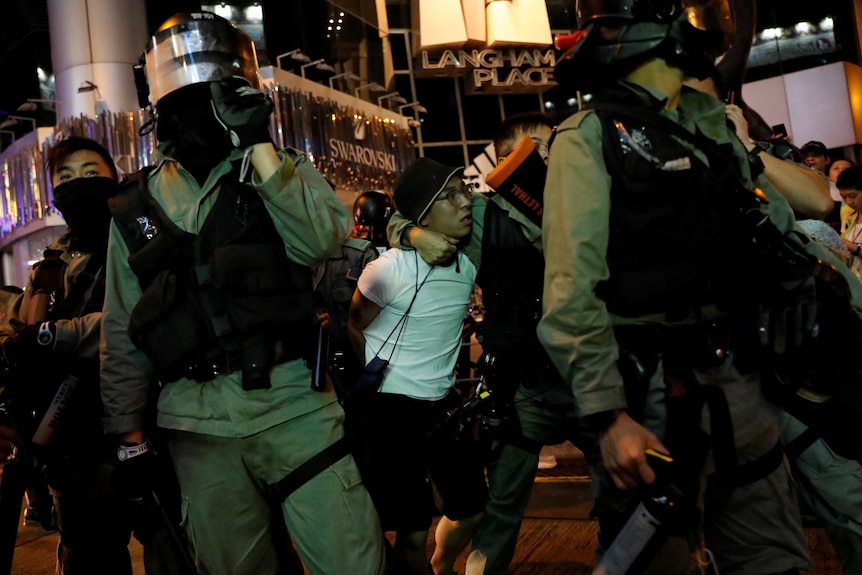 A protester in a white shirt and glasses is dragged away by a group of police in riot gear