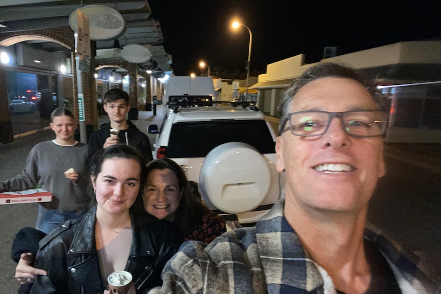 A middle-aged man takes a selfie with his children and wife on a street at night.