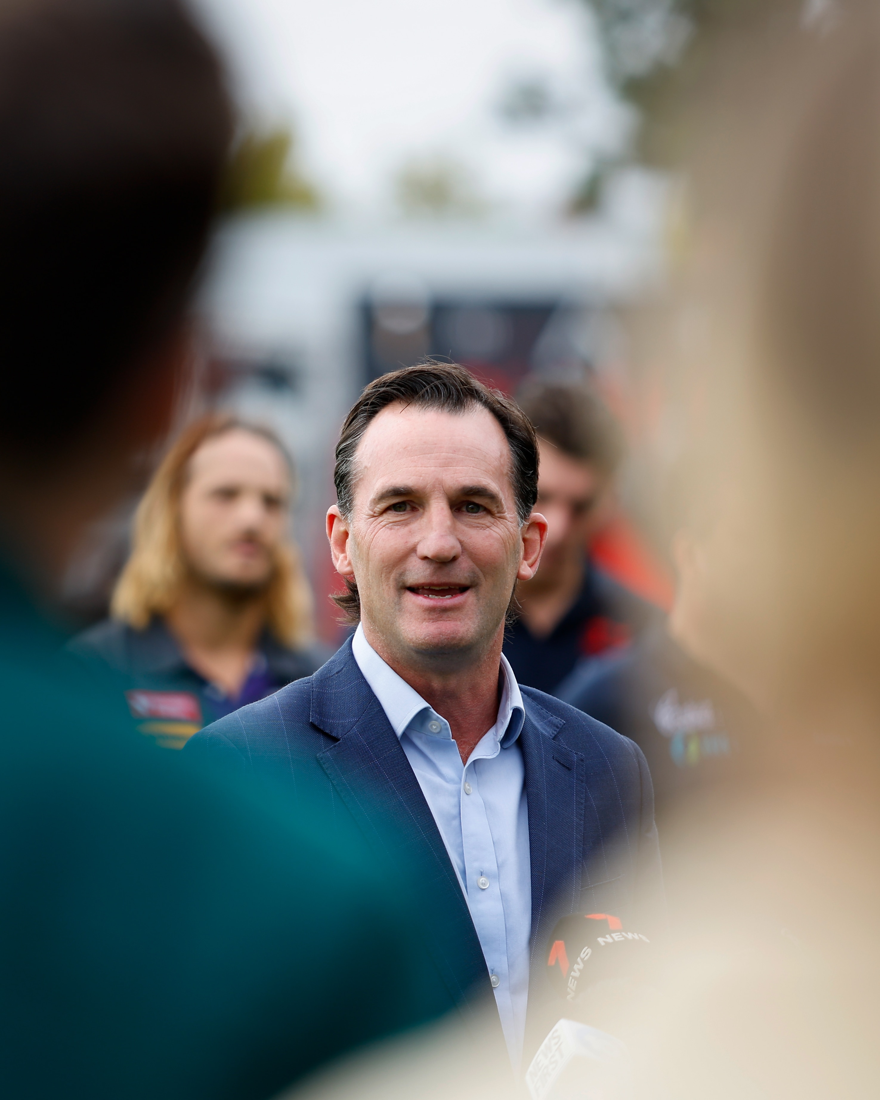 O executivo da AFL, Andrew Dillon, fala à mídia, emoldurado por figuras borradas em primeiro plano.