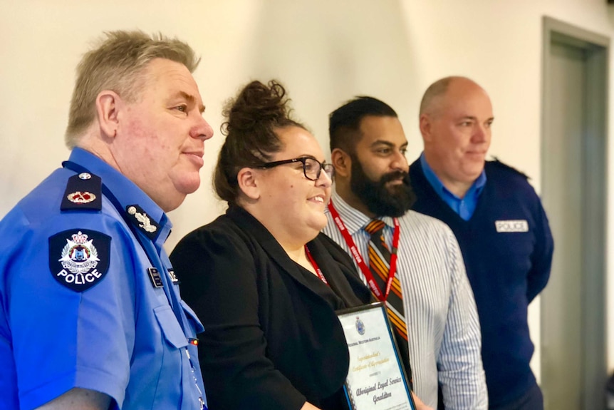 Four people standing in front of a white wall in the Geraldton police station holding an award.