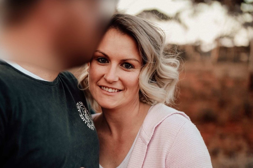 A blonde woman stares at the camera, with a red dirt background.