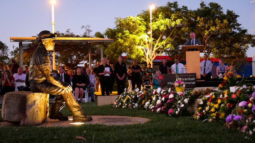 A man speaks at a lectern at dusk in front of a large crowd gathered around a statue of a coal miner.