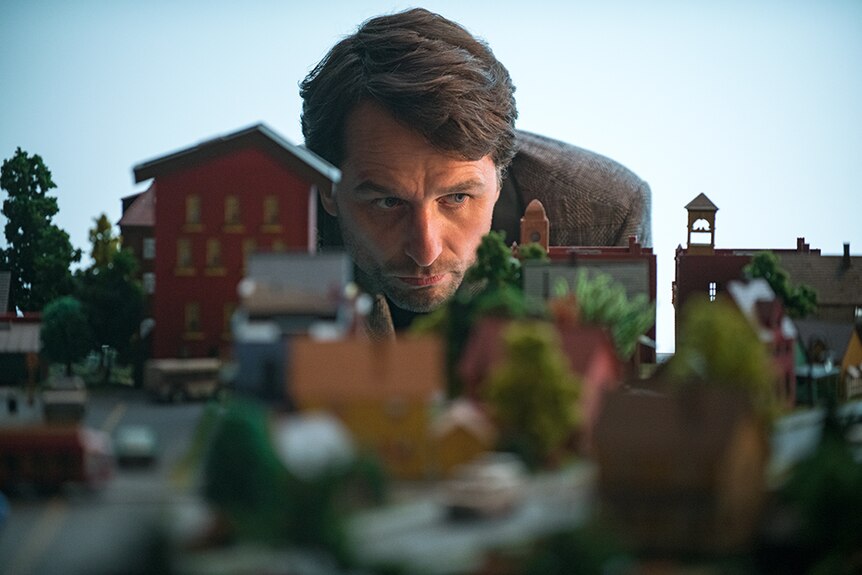 A man with focused expression and short brown hair bends over to inspect miniature model featuring buildings and trees.