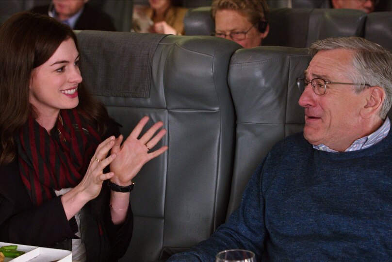 A young woman with brown hair gesticulates as she converses with an older bespectacled man seated next to her on airplane.