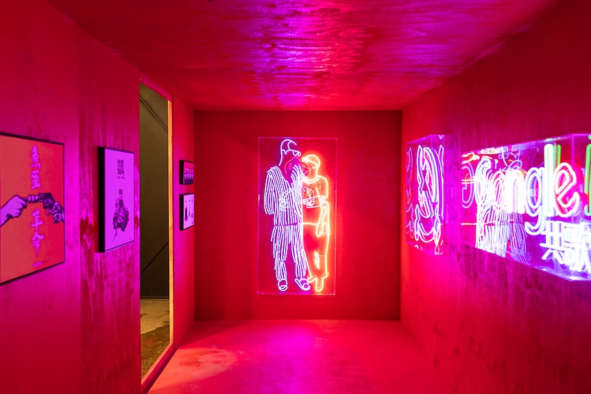 A neon light sculpture of two figures embracing illuminates a small gallery space with suspended artworks.