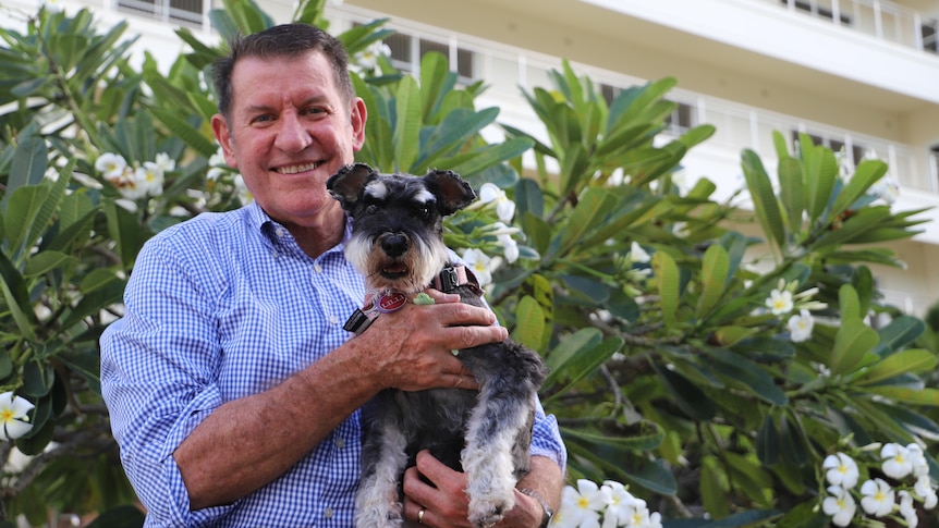 A man holding a small dog looking at the camera and smiling in front of trees and a unit building.