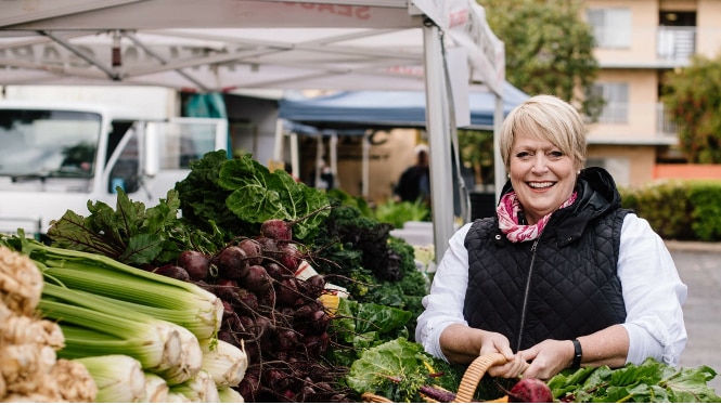 blonde haired woman in white top and black vest holding a basket of vegetables in front of celery and beetroots