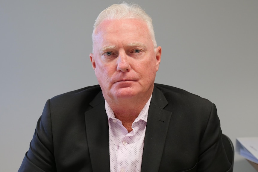 A man in a black suit sitting at a desk looking solemn.